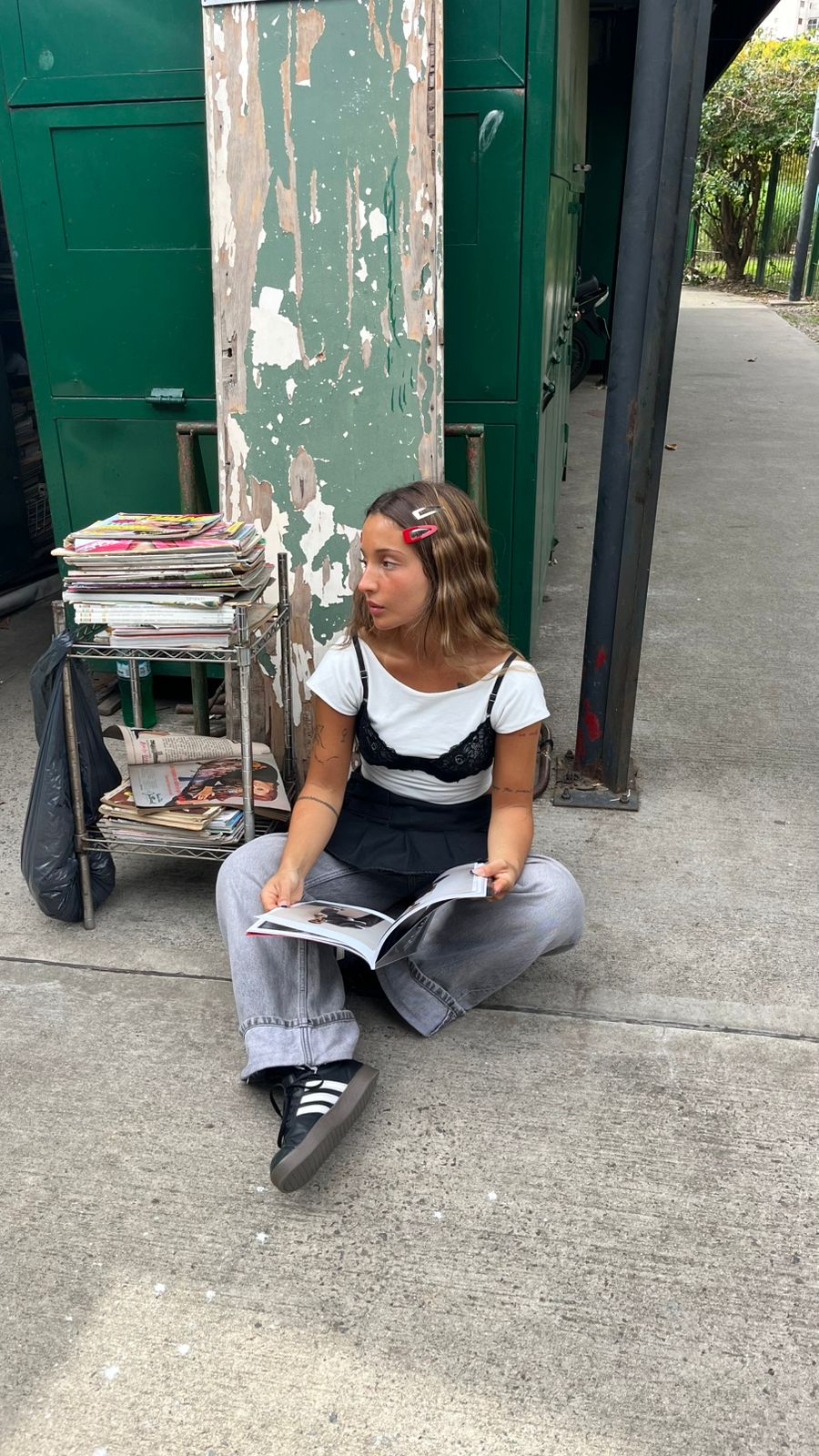 Camila sitting on the street in a lace bra over a white tee, baggy jeans and Adidas Sambas reading a magazine